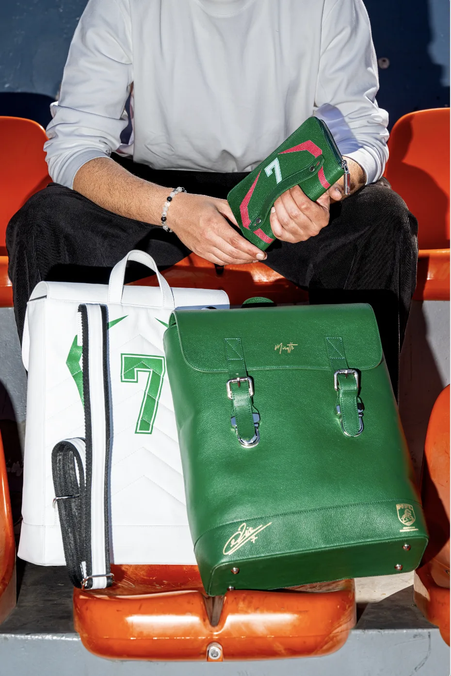 La collection JZ7 dans les gradins d&rsquo;un stade : les backpacks Mousafir vert et blanc, le porte-passeport Jawal tenu en main